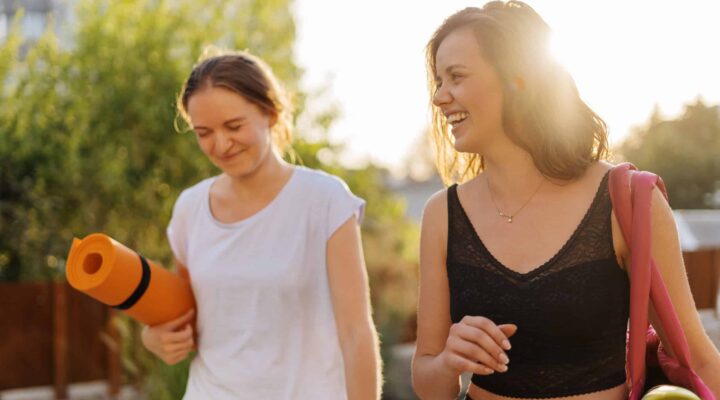 Dos mujeres jóvenes con esterillas de yoga caminando al aire libre para practicar ejercicio físico y mantener una vida saludable.
