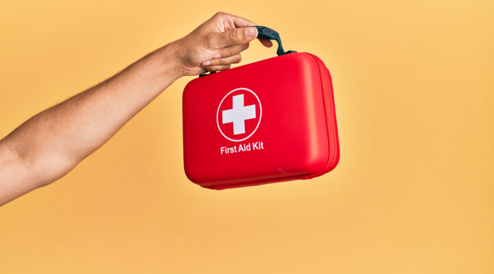 Mano sosteniendo un botiquín de primeros auxilios rojo sobre fondo amarillo.
